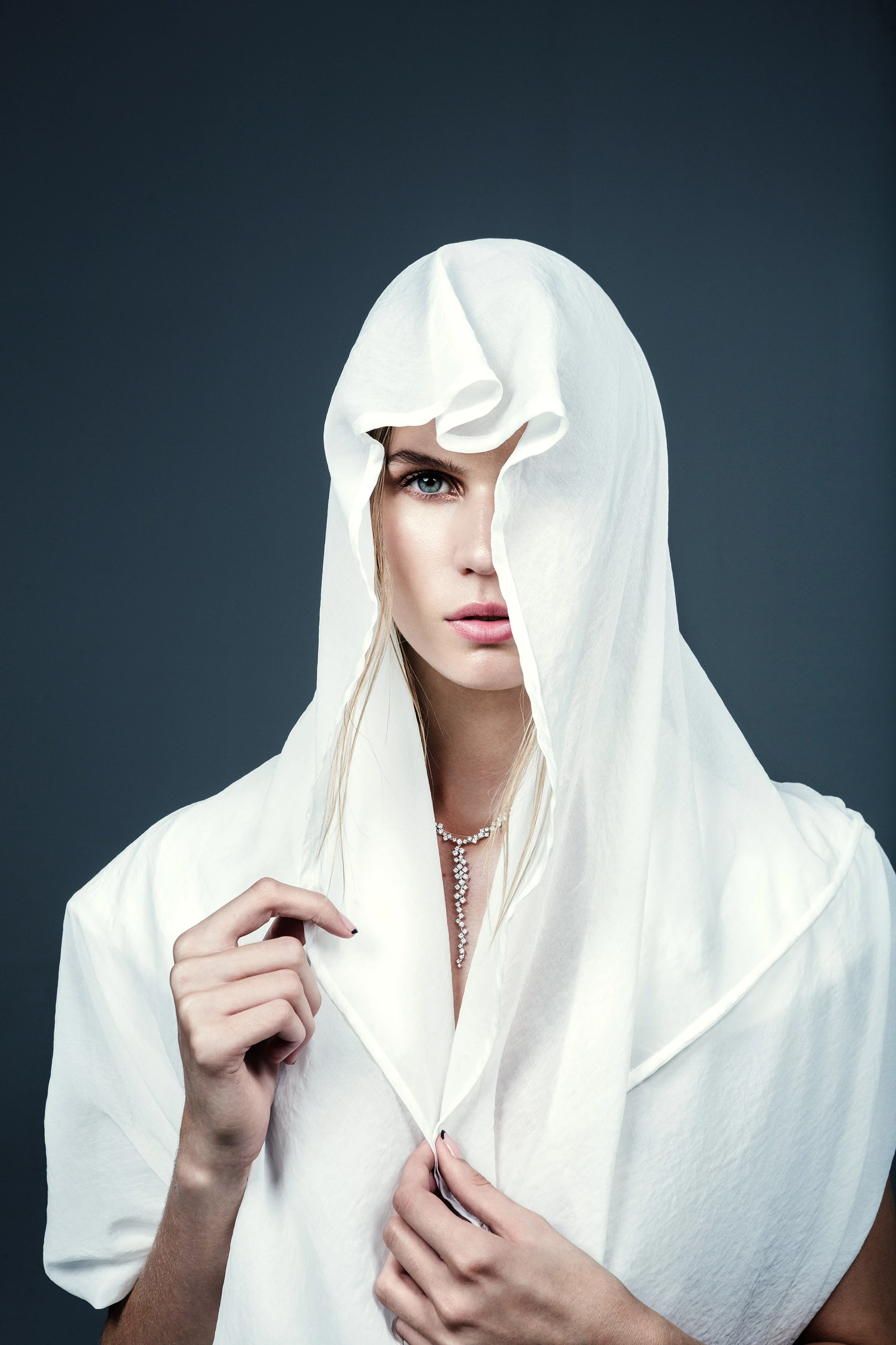 Portrait of a woman with intense blue eyes draped in a white veil against a muted blue background, wearing a delicate pearl necklace.