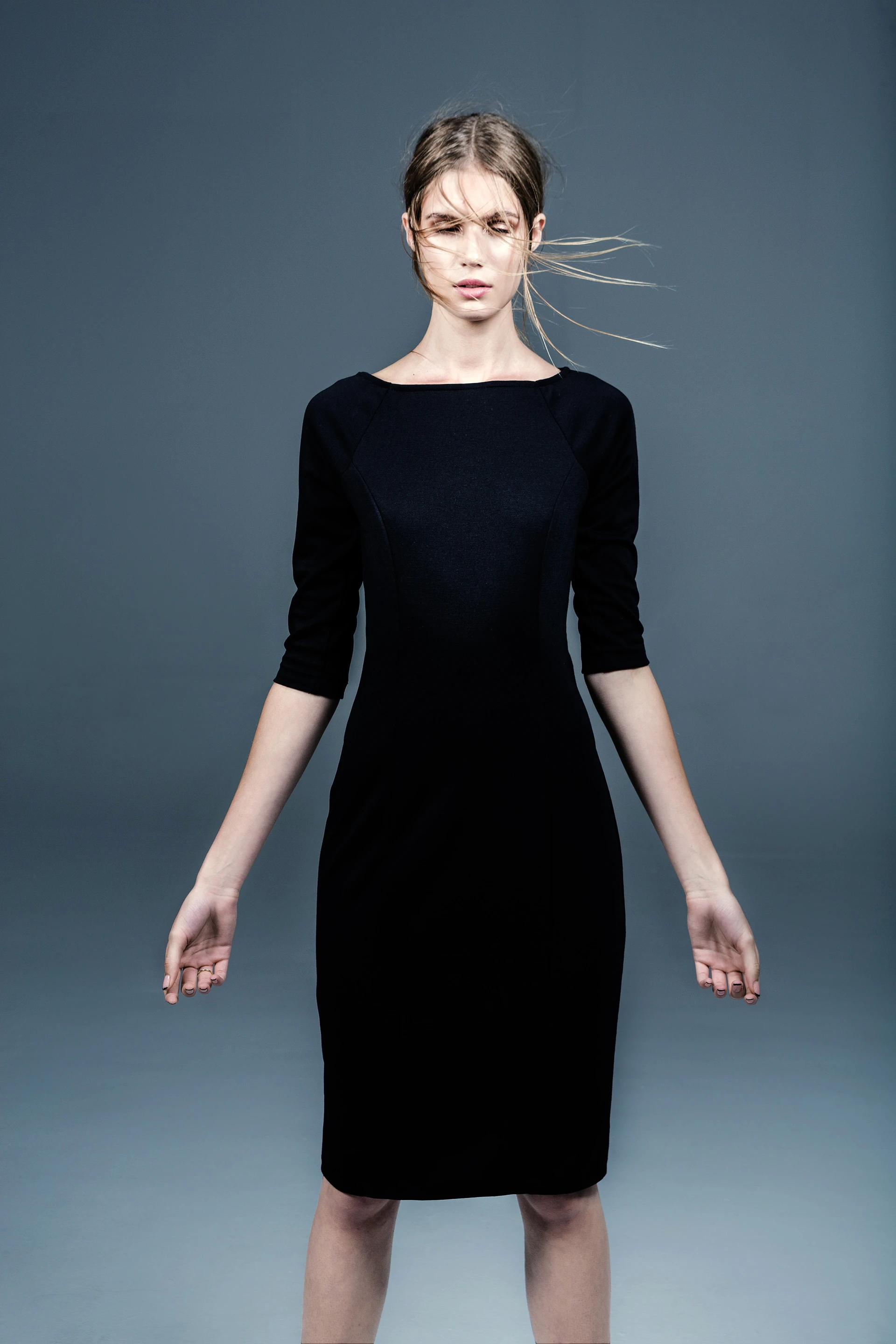 Woman in a fitted black dress, with strands of her hair blowing across her face, against a gray-blue backdrop.